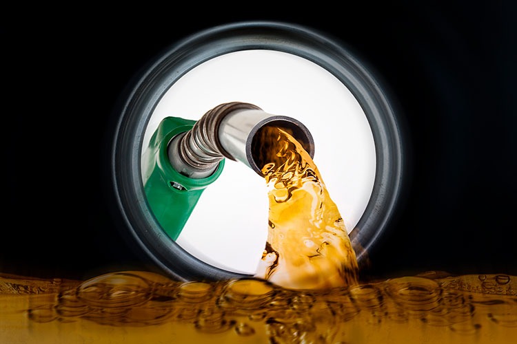 Primer plano de una boquilla de combustible con mango verde que vierte un líquido de color amarillo-marrón —probablemente gasolina o gasóleo— en la abertura redonda de un depósito —una escena cotidiana para quienes dependen de la carretera para su trabajo—, mientras el líquido salpica y forma burbujas en el fondo, reflejando el precio del combustible, siempre cambiante.